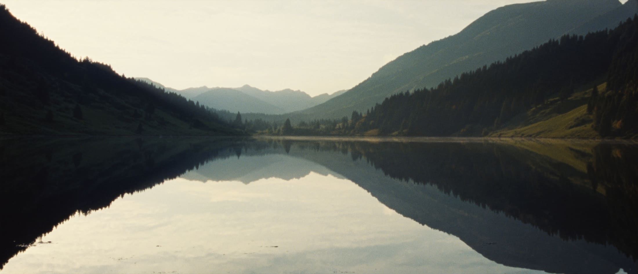 A still mountain lake at first light, with forested slopes mirrored on the water