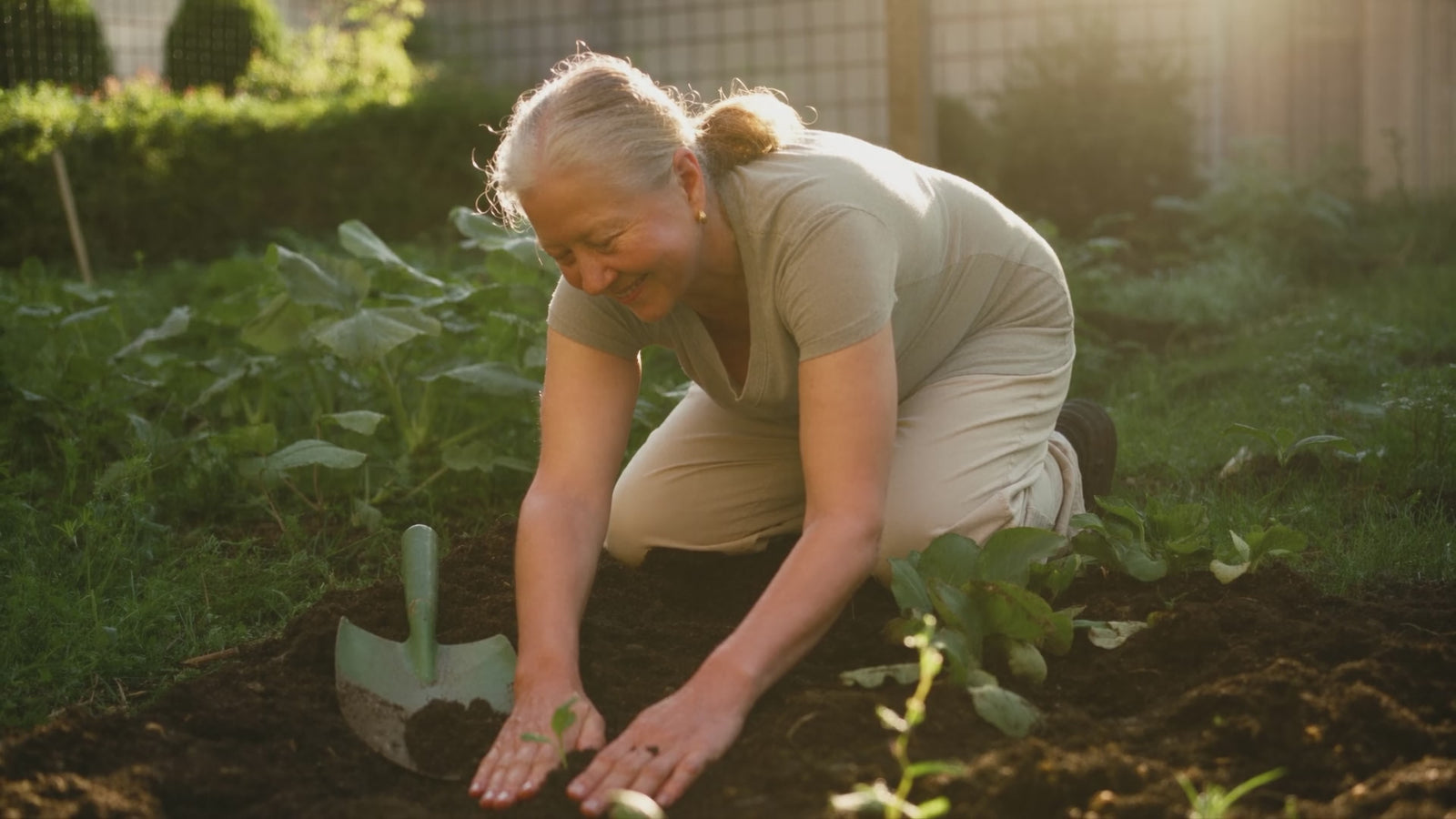 An older woman planting seedlings in a sunlit vegetable garden, smiling, in gentle motion