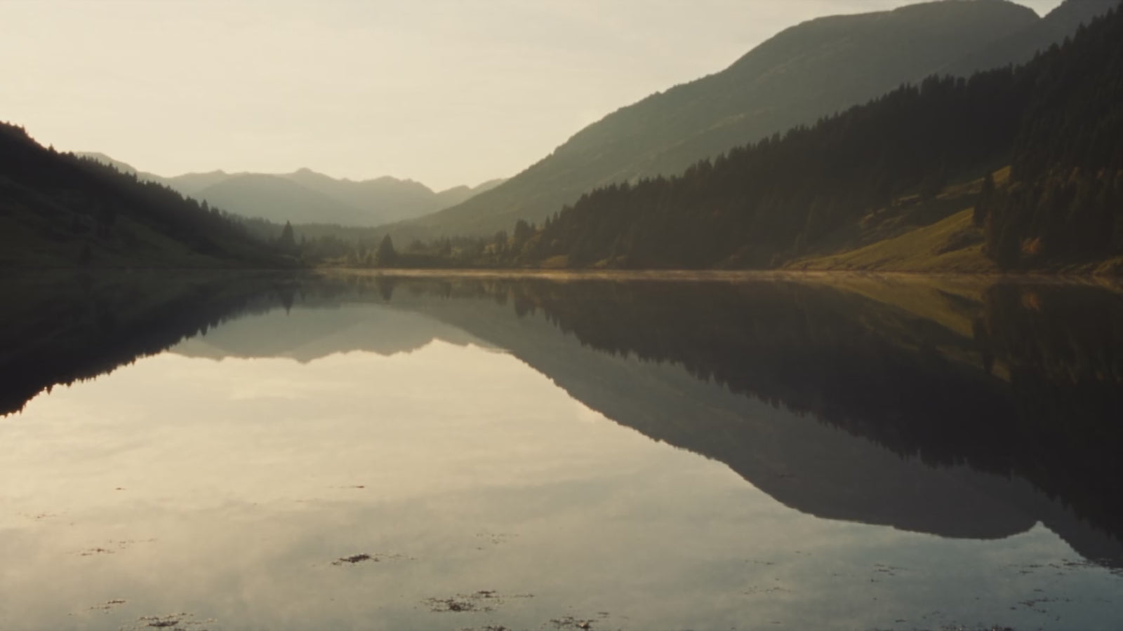 A still mountain lake at first light, mist drifting over the water, forested slopes mirrored on the surface