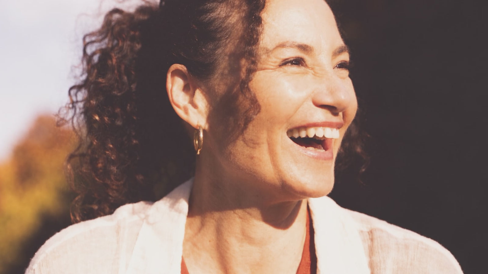 A woman laughing in autumn sunlight, one hand resting on her chest, in gentle motion