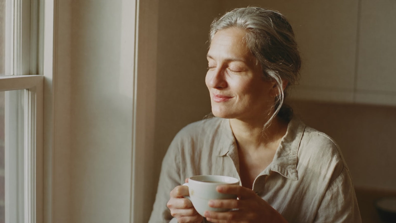 A woman at a sunlit kitchen window in the morning, holding a mug of tea, in gentle motion